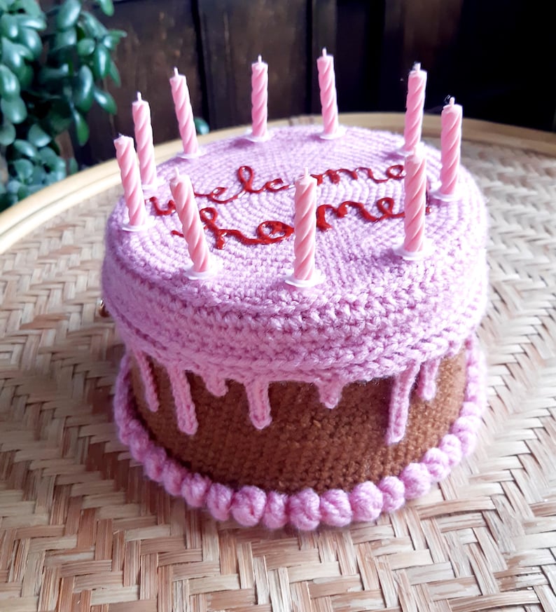 May include: A crocheted cake with a brown base and a pink top, adorned with pink candles. The top of the cake has the words "welcome home" embroidered in red. The cake sits on a woven tray.