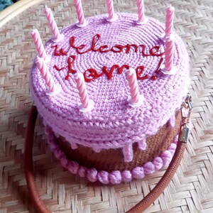May include: A crocheted cake-shaped handbag in shades of pink and brown, adorned with pink candles. The words "welcome home!" are embroidered in red on the top. The bag has a brown strap and a gold-colored zipper.