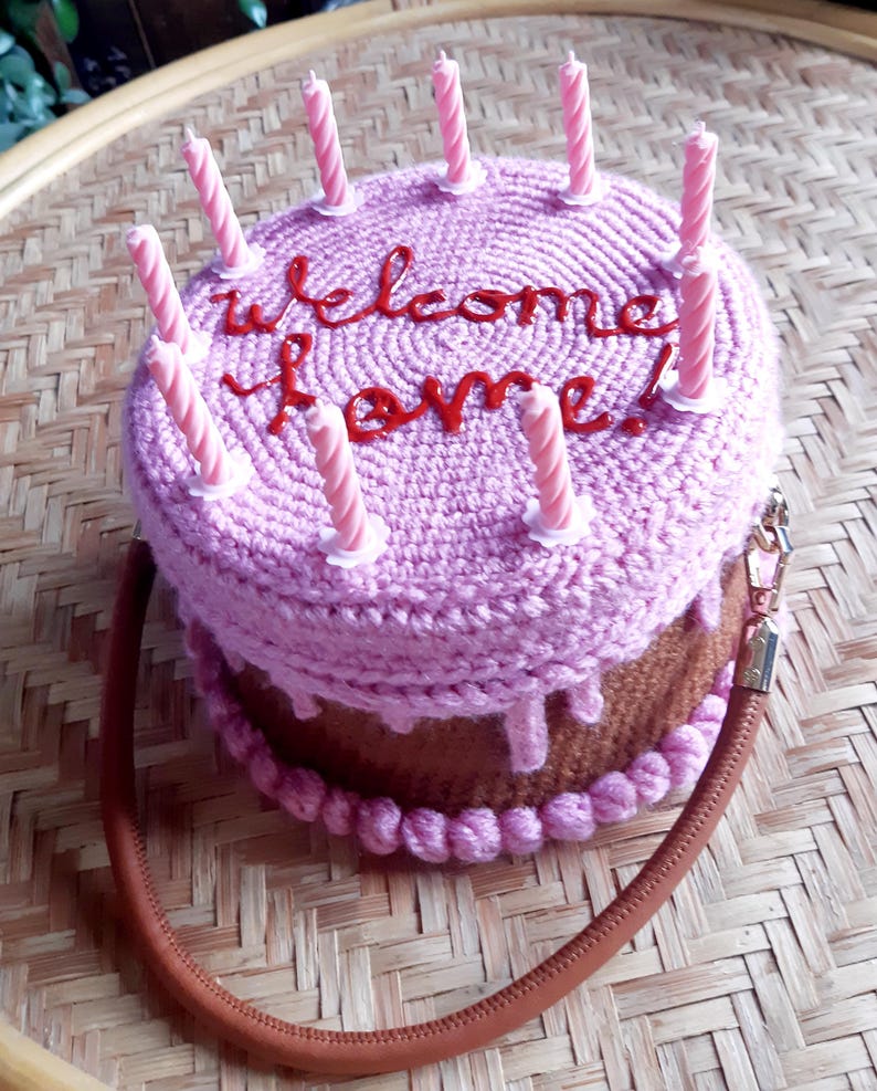 May include: A crocheted cake-shaped handbag in shades of pink and brown, adorned with pink candles. The words "welcome home!" are embroidered in red on the top. The bag has a brown strap and a gold-colored zipper.