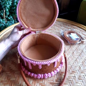 May include: A round, open handbag resembling a cake, with a brown exterior and pink frosting details. The bag has a brown handle and a matching strap. The interior is lined with a soft, brown fabric. The zipper is gold-toned.