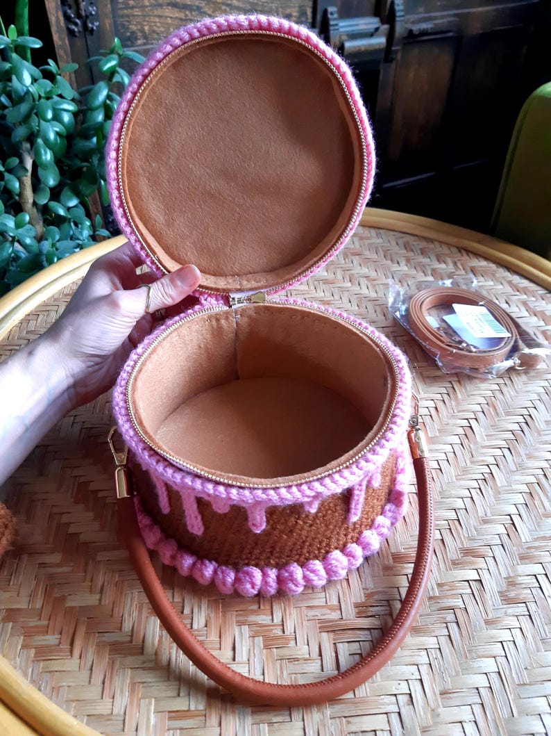 May include: A round, open handbag resembling a cake, with a brown exterior and pink frosting details. The bag has a brown handle and a matching strap. The interior is lined with a soft, brown fabric. The zipper is gold-toned.