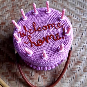 May include: A crocheted purple handbag shaped like a cake, with the words "Welcome home!" in red script. The cake has pink candles and a brown strap. The bag is sitting on a woven surface.
