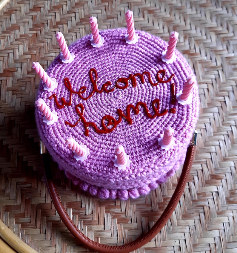 May include: A crocheted purple handbag shaped like a cake, with the words "Welcome home!" in red script. The cake has pink candles and a brown strap. The bag is sitting on a woven surface.