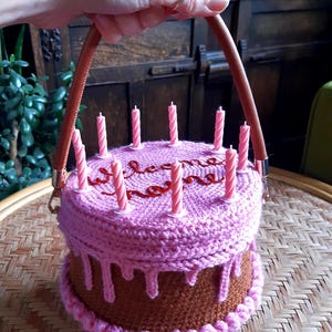 May include: A crocheted cake-shaped handbag with brown and pink yarn. The bag has a brown base, pink frosting, and pink candles. The handle is brown leather. The word "welcome home" is embroidered on top.