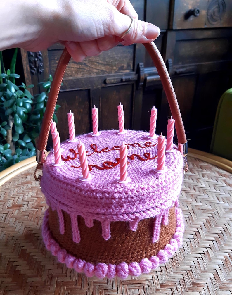 May include: A crocheted cake-shaped handbag with brown and pink yarn. The bag has a brown base, pink frosting, and pink candles. The handle is brown leather. The word "welcome home" is embroidered on top.