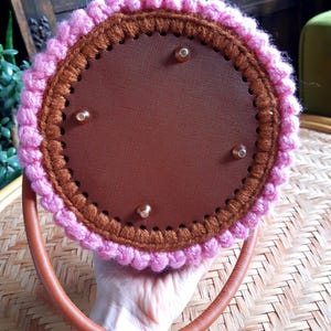 May include: A round, handmade handbag with a brown leather handle. The bag features a brown interior and is trimmed with pink and brown crochet. The bag has four gold-colored rivets.