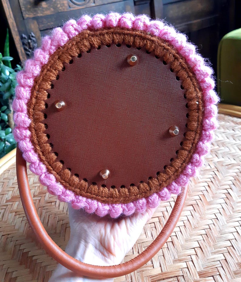 May include: A round, handmade handbag with a brown leather handle. The bag features a brown interior and is trimmed with pink and brown crochet. The bag has four gold-colored rivets.