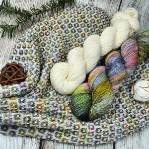 May include: An assortment of colorful yarn skeins and a knitted fabric piece. The yarn includes white, multicolored, and variegated options. The knitted item features a geometric pattern with various colors. The arrangement is on a wooden surface.