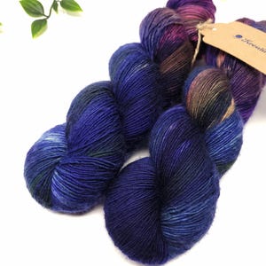 Könnte beinhalten: Zwei Knäuel Garn in einem tiefen Blau-, Lila- und Grünton. Das Garn ist handgefärbt und hat ein melierter Muster. Die Knäuel sind mit "Kreativ" beschriftet.