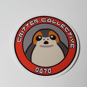 May include: A round sticker with a red border and a white background. The sticker features a cartoon image of a Porg, a small, furry creature from the Star Wars universe. The text "Critter Collective" is written in a circular pattern around the image, and the text "0070" is written below the image.