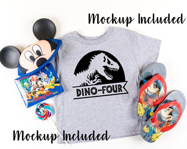 May include: A gray t-shirt with a black dinosaur graphic and the text "Dino-Four". The shirt is on a white surface with a Mickey Mouse hat, a book, a lollipop, and flip-flops.