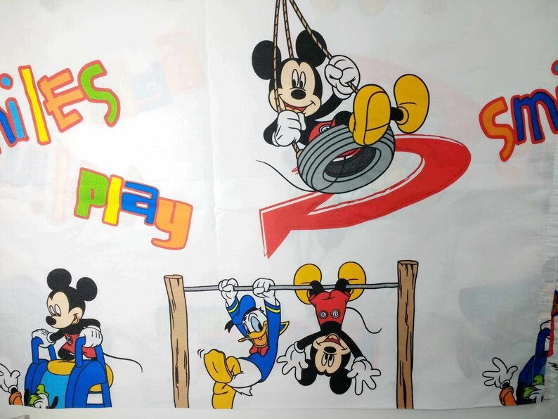 K&ouml;nnte beinhalten: Ein wei&szlig;er Stoff mit einem bunten Druck, der Mickey Mouse und Donald Duck zeigt. Der Druck enth&auml;lt die W&ouml;rter "Smiles Play" und "Smile".