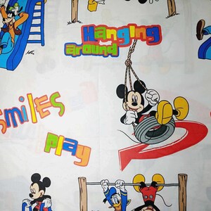 K&ouml;nnte beinhalten: Wei&szlig;er Stoff mit einem bunten Cartoon-Druck mit Mickey Mouse und Goofy. Der Druck enth&auml;lt die W&ouml;rter "Hanging Around", "Smiles" und "Play".