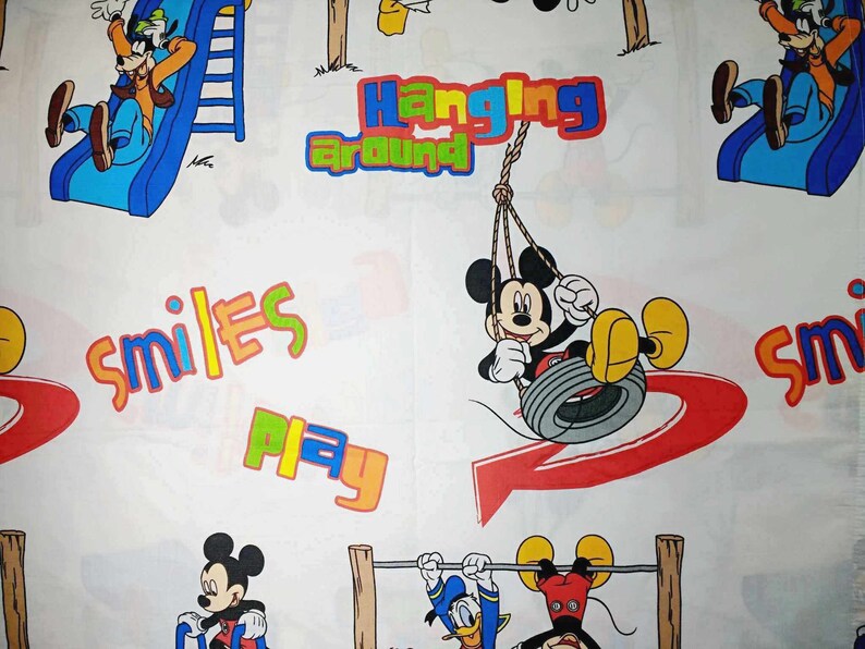 K&ouml;nnte beinhalten: Wei&szlig;er Stoff mit einem bunten Cartoon-Druck mit Mickey Mouse und Goofy. Der Druck enth&auml;lt die W&ouml;rter "Hanging Around", "Smiles" und "Play".