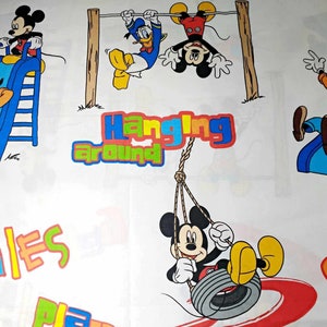 K&ouml;nnte beinhalten: Ein wei&szlig;er Stoff mit einem farbenfrohen Cartoon-Design mit Mickey Mouse und seinen Freunden. Das Design zeigt Mickey Mouse auf einer Schaukel, Mickey Mouse auf einer Rutsche, Mickey Mouse, der an einer Stange h&auml;ngt, und Goofy auf einer Rutsche. Der Text "Hanging Around" und "Smiles" ist ebenfalls im Design enthalten.