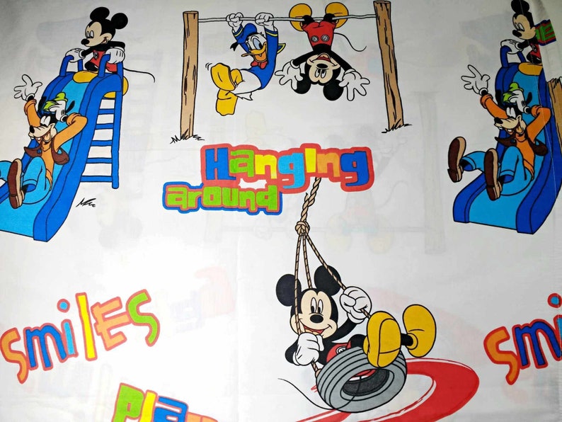 K&ouml;nnte beinhalten: Ein wei&szlig;er Stoff mit einem farbenfrohen Cartoon-Design mit Mickey Mouse und seinen Freunden. Das Design zeigt Mickey Mouse auf einer Schaukel, Mickey Mouse auf einer Rutsche, Mickey Mouse, der an einer Stange h&auml;ngt, und Goofy auf einer Rutsche. Der Text "Hanging Around" und "Smiles" ist ebenfalls im Design enthalten.