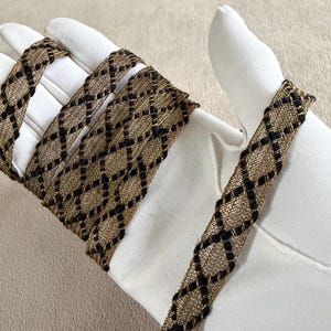 May include: Gold and black decorative ribbon with a diamond pattern. The ribbon is woven with a metallic gold thread and black accents. The ribbon is shown coiled on a white surface.
