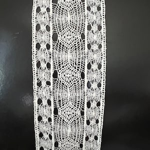 May include: Close-up of a white lace ribbon with an intricate, openwork design. The lace features a repeating pattern of circular and oval shapes, creating a delicate and textured appearance. The ribbon is against a dark background.