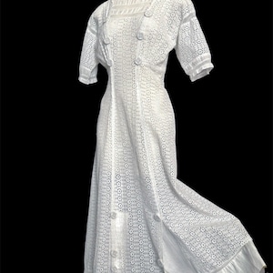 May include: A white, vintage-style dress with a high neckline, short sleeves, and a button-up front. The dress features intricate openwork detailing and a flowing skirt, perfect for a special occasion. The dress is displayed on a mannequin against a black background.