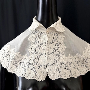 May include: An antique white lace collar with a delicate floral design. The collar is buttoned in the front and has a scalloped edge. The lace is intricately detailed, creating a vintage aesthetic.