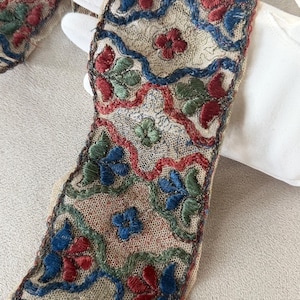 May include: Close-up of an embroidered textile with a floral pattern. The design features red, blue, and green flowers and vines on a beige background. The embroidery is detailed and intricate, showcasing a vintage aesthetic.