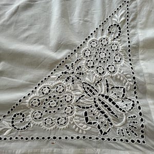 May include: White fabric with a detailed embroidered design in a triangular shape. The embroidery features floral and geometric patterns, with small circular cutouts along the edges. The fabric has a slight sheen.