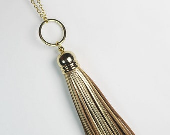 May include: A gold-tone chain necklace featuring a circular pendant attached to a long, multi-strand tassel. The tassel consists of alternating strips of metallic gold and matte brown material, secured by a polished gold-tone cap at the top.