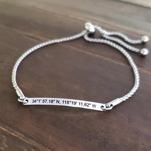 K&ouml;nnte beinhalten: Ein verstellbares Armband aus Silber mit einem silbernen Anh&auml;nger, auf dem die Koordinaten "34&deg;1' 57.18" N, 118&deg;19' 11.62" W" eingraviert sind.