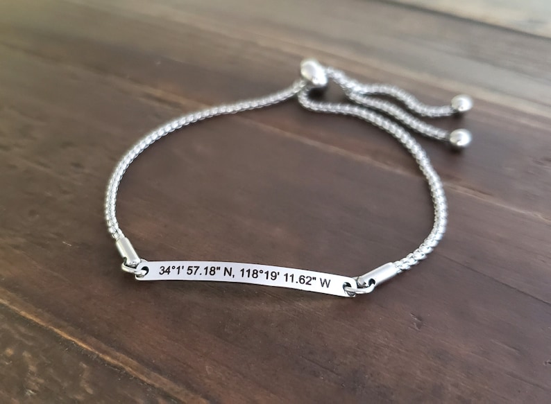 K&ouml;nnte beinhalten: Ein verstellbares Armband aus Silber mit einem silbernen Anh&auml;nger, auf dem die Koordinaten "34&deg;1' 57.18" N, 118&deg;19' 11.62" W" eingraviert sind.