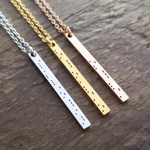 Puede incluir: Tres collares de plata, oro y oro rosa con colgantes de barra grabados con puntos Braille. Los collares están sobre una superficie de madera.