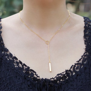 May include: A delicate gold-colored necklace featuring a small circle and a vertical bar pendant. The necklace is worn against a dark blue lace top, creating a contrast. The chain is fine and elegant.