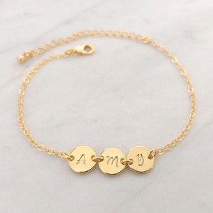 Könnte beinhalten: Ein goldenes Kettenarmband mit drei runden Anhängern, die jeweils mit einem Buchstaben graviert sind und den Namen "AMY" ergeben.
