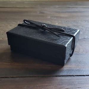 K&ouml;nnte beinhalten: Eine schwarze Geschenkbox mit einem schwarzen Band, das zu einer Schleife gebunden ist.