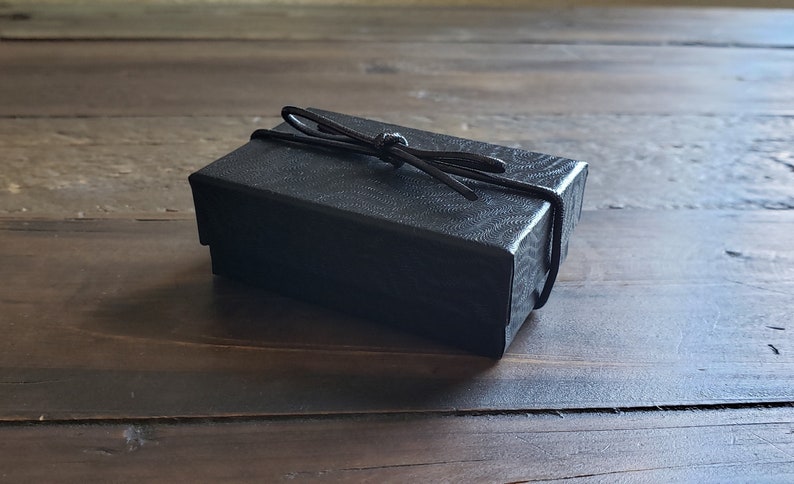 K&ouml;nnte beinhalten: Eine schwarze Geschenkbox mit einem schwarzen Band, das zu einer Schleife gebunden ist.