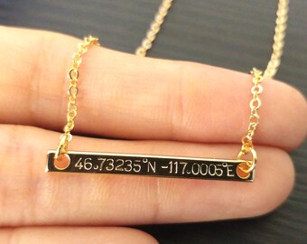 May include: A gold-colored bar necklace with a delicate chain. The bar is engraved with the coordinates 46.73235°N -117.0005°E. The necklace is held in a person's hand.