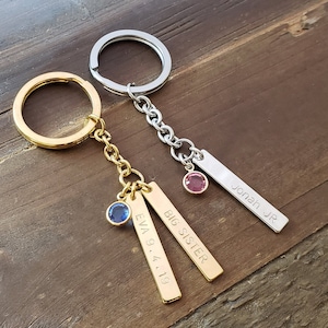 May include: Two gold and silver keychains with engraved tags. The gold keychain has a blue gem and the tags read "BIG SISTER" and "EVA 9.4.19". The silver keychain has a pink gem and the tag reads "JONAH JR".