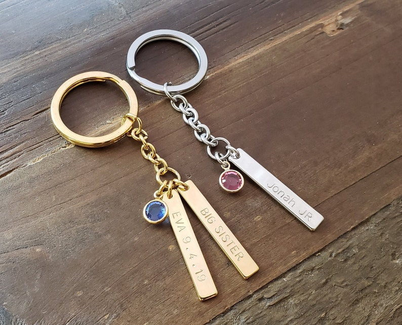 May include: Two gold and silver keychains with engraved tags. The gold keychain has a blue gem and the tags read "BIG SISTER" and "EVA 9.4.19". The silver keychain has a pink gem and the tag reads "JONAH JR".