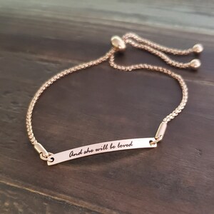 K&ouml;nnte beinhalten: Ein verstellbares Armband aus Ros&eacute;gold mit einer zarten Kette und einem kleinen rechteckigen Anh&auml;nger mit der Aufschrift "And she will be loved".
