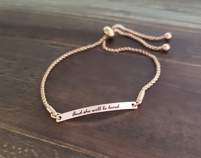 K&ouml;nnte beinhalten: Ein verstellbares Armband aus Ros&eacute;gold mit einer zarten Kette und einem kleinen rechteckigen Anh&auml;nger mit der Aufschrift "And she will be loved".