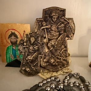 May include: Bronze-colored religious statues of Saint Michael the Archangel, one large and one small, with a rosary in the foreground and a painting of a saint in the background. The statues depict Saint Michael with a sword and shield.