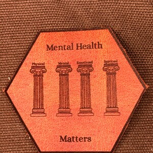 May include: A hexagonal, orange leather patch with the words "Mental Health Matters." Four columns are below the words, each labeled with a different aspect of health: Physical, Mental, Emotional, and Spiritual. The patch is on a brown, textured surface.