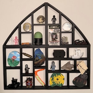 Puede incluir: Una estantería negra con forma de casa llena de varios objetos pequeños. Los artículos incluyen monedas, figuritas, rocas pintadas, una figura de acción de Boba Fett y un pequeño barco. La estantería está montada en una pared blanca.