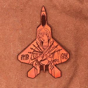 May include: A laser-cut patch in the shape of a fighter jet, featuring a religious icon with multiple swords. The patch is orange and brown, with the number 93 visible. The background is a textured, brown fabric.