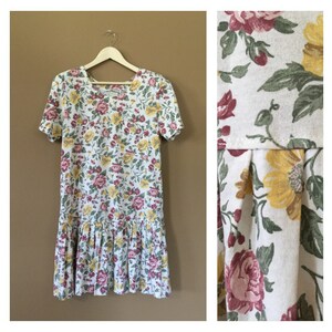 K&ouml;nnte beinhalten: Ein wei&szlig;es Kleid mit einem Blumendruck in Rosa-, Gelb- und Gr&uuml;nt&ouml;nen. Das Kleid hat kurze &Auml;rmel und einen R&uuml;schen-Saum.