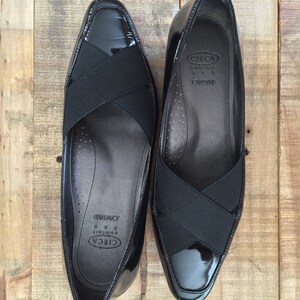 May include: A pair of black patent leather shoes with a black elastic strap across the vamp. The shoes have a pointed toe and a low heel. The brand name "CIRCA" and the text "Comfort 8 1/2" are printed on the inside of the shoes.