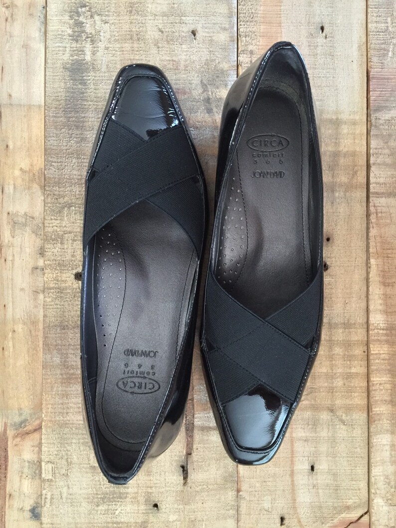 May include: A pair of black patent leather shoes with a black elastic strap across the vamp. The shoes have a pointed toe and a low heel. The brand name "CIRCA" and the text "Comfort 8 1/2" are printed on the inside of the shoes.