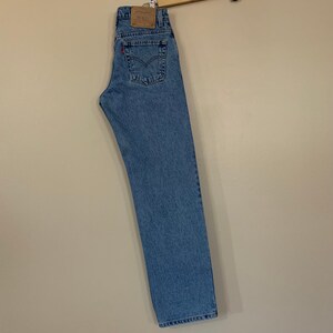 May include: A pair of light blue denim jeans hanging on a wooden hanger. The jeans have a brown leather patch with the Levi's logo on the back.
