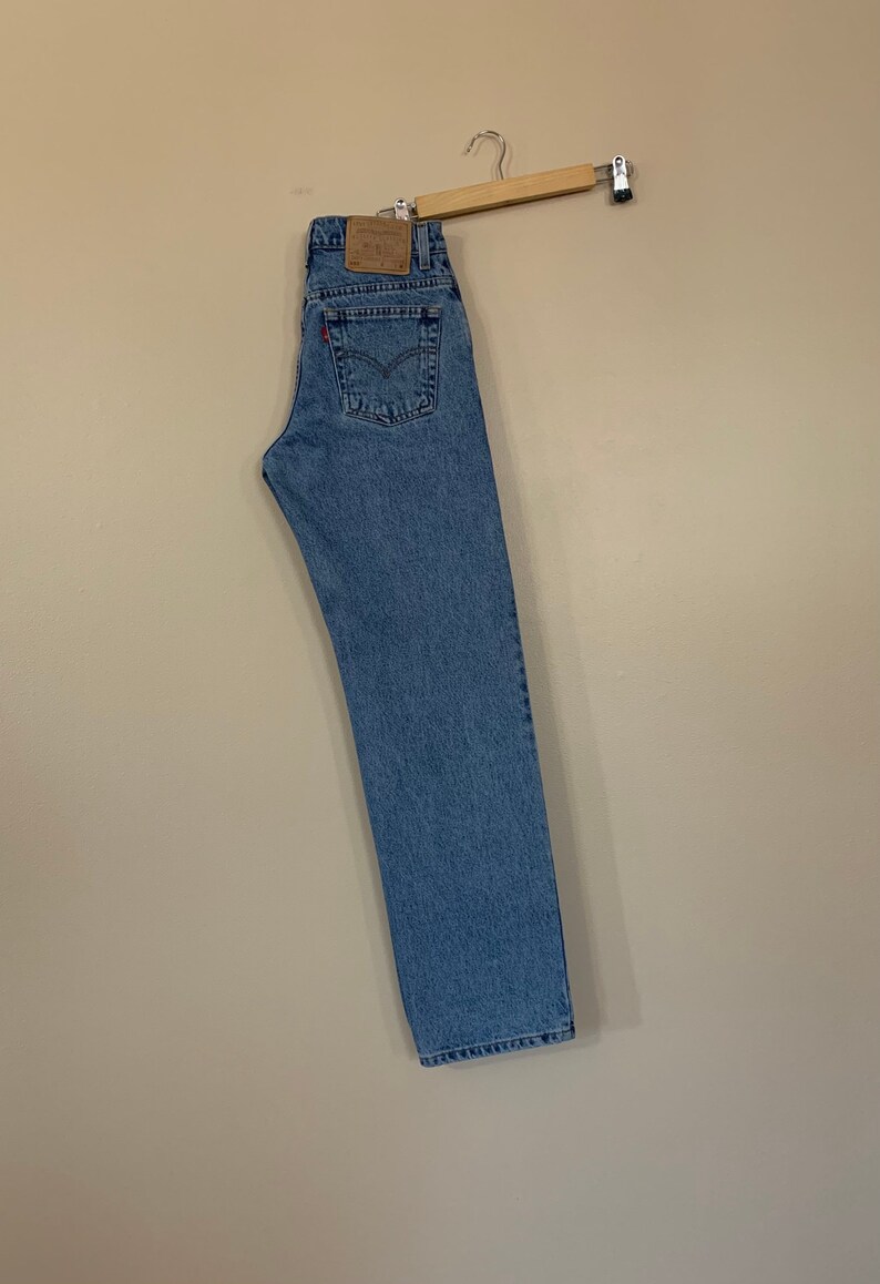 May include: A pair of light blue denim jeans hanging on a wooden hanger. The jeans have a brown leather patch with the Levi's logo on the back.