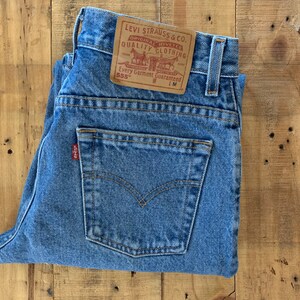 May include: Light blue denim jeans with a classic five-pocket design. The jeans feature a leather patch with the Levi Strauss & Co. logo and the text "ORIGINAL RIVETED QUALITY CLOTHING". A small red tag is visible on the back pocket.