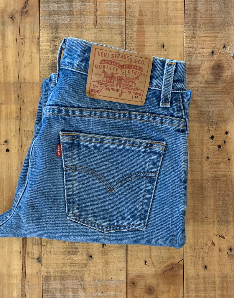 May include: Light blue denim jeans with a classic five-pocket design. The jeans feature a leather patch with the Levi Strauss & Co. logo and the text "ORIGINAL RIVETED QUALITY CLOTHING". A small red tag is visible on the back pocket.